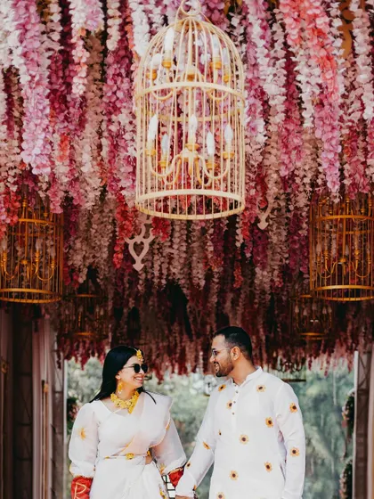 The couple stands under a canopy of hanging pink and white wisteria, with birdcage chandeliers adding a whimsical touch. This shot captures the enchanting and detailed decor of their Haldi ceremony.