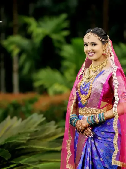 A full-length bridal portrait showing a bride in her stunning blue and pink saree, complete with a veil, in a beautiful garden setting.