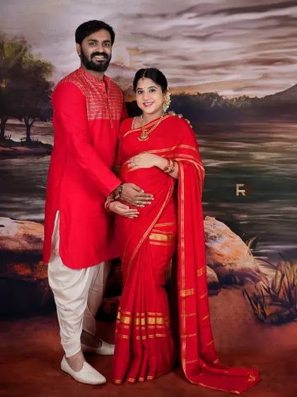 A classic couple's portrait in matching red traditional outfits. The dad-to-be gently holds the baby bump, a simple and powerful expression of his love.