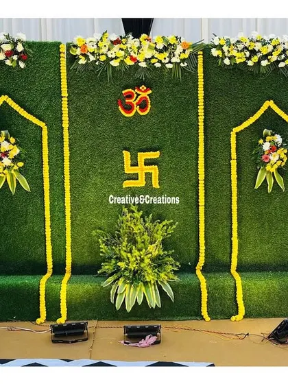 A simple and auspicious backdrop for a religious ceremony. The green turf wall is decorated with 'Om' and 'Swastik' symbols made from marigolds.