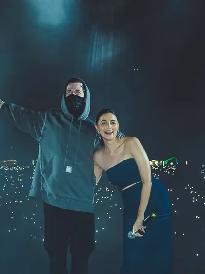 Alan Walker and Alia Bhatt pose for a photo on stage, with a sea of phone lights from the audience behind them.