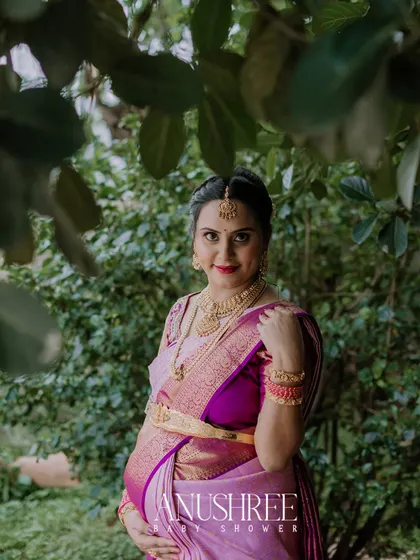 A beautiful outdoor portrait from a traditional baby shower photoshoot. The mother-to-be is framed by lush greenery, which makes the vibrant colors of her saree and her happy expression stand out.
