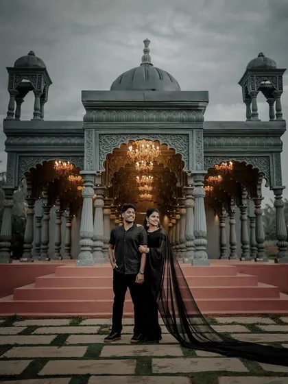 A stunning wide shot of the couple against the dramatic architecture of Shire Studio, with the bride's long black saree creating a beautiful silhouette.