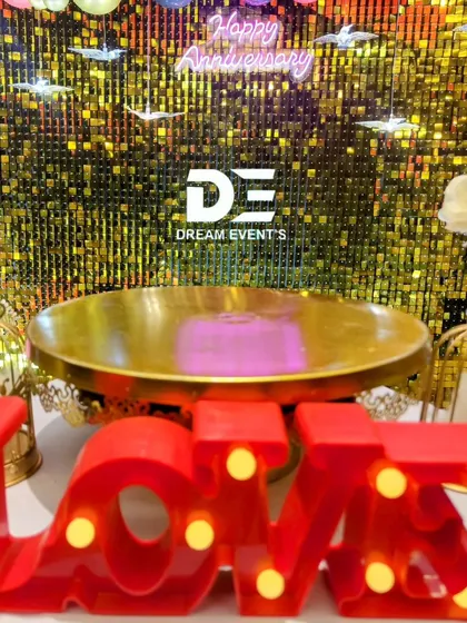 A detailed shot of an anniversary setup, showing a 'LOVE' marquee sign and a golden cake stand in front of a shimmering sequin backdrop.