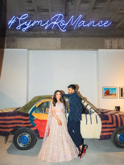 The couple poses in front of a quirky, carpet-covered vintage car and a custom neon sign, adding a fun, modern art twist to their Sangeet night.