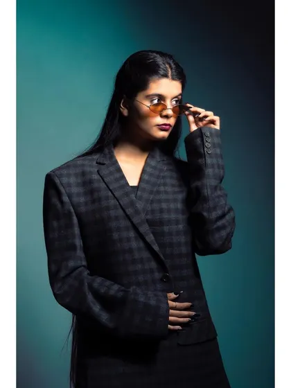 A studio shot of a model in a plaid blazer, adjusting her sunglasses. The teal background and professional lighting create a polished, high-fashion look suitable for a brand's lookbook or magazine feature.