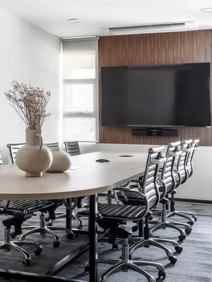 A modern conference room where natural wood and clean lines meet. The large screen is integrated into a wood-paneled feature wall, and a simple vase with dried flowers adds a soft, organic touch to the professional setting.