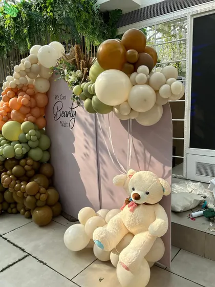 A cuddly teddy bear sits on a cloud of white balloons, holding a smaller balloon bunch. This floating effect adds a touch of whimsy to the earthy-toned setup.