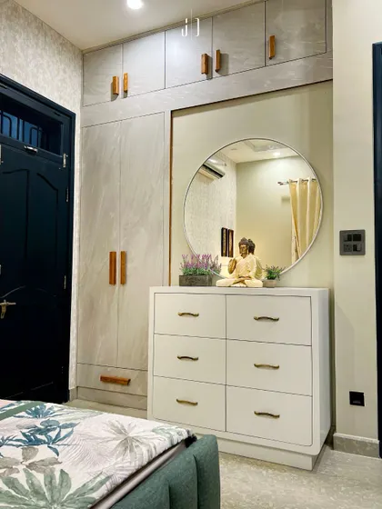 A functional and stylish bedroom corner with a modern wardrobe and a chest of drawers that doubles as a dressing table. The round mirror and Buddha statue add a sense of calm.