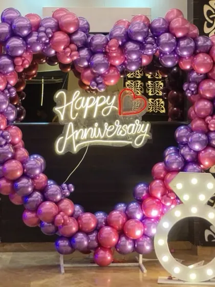 A romantic anniversary decoration featuring a heart-shaped balloon arch in purple and pink. A "Happy Anniversary" neon sign and a light-up ring prop complete this perfect photo spot.