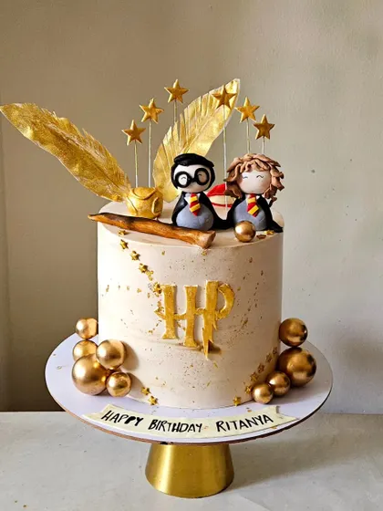 A magical Harry Potter cake for a fan's birthday. This design features adorable, handcrafted figurines of Harry and Hermione, a golden snitch, and a wand, all set against a backdrop of golden stars and feathers.