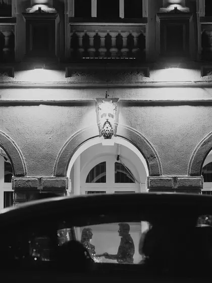 A secret moment captured through a car window. This black and white photograph has a voyeuristic, cinematic feel, framing the couple against the beautifully lit architecture in the background for a timeless pre-wedding shot.