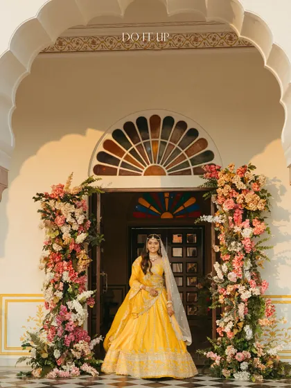 The bride, framed by a magnificent floral installation at the entrance of the heritage venue in Jaipur. The design uses a palette of pinks, peaches, and whites to create a soft yet grand welcome.