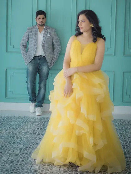 A stylish studio shot where the mother-to-be, radiant in her yellow gown, is the focus. Her partner stands in the background, creating a sense of depth and a modern, artistic composition for this maternity portrait.
