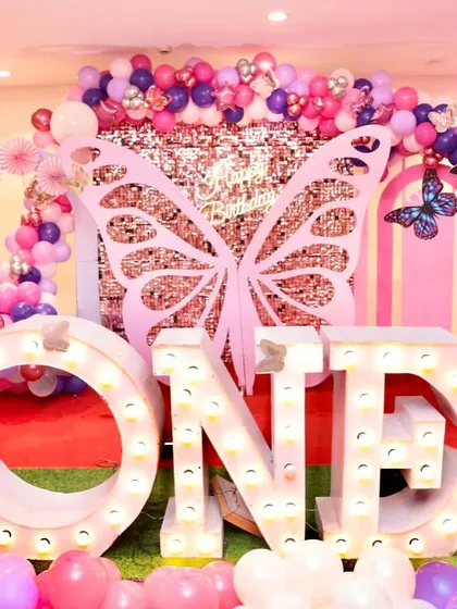A first birthday party with a pink and purple butterfly theme. The stage is set with large "ONE" marquee letters, a sequin backdrop, and butterfly wing props.
