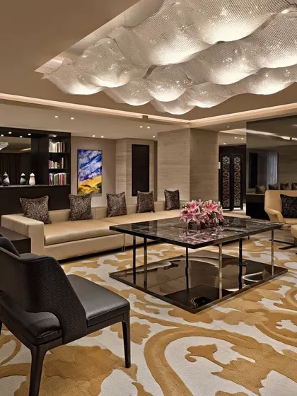 A luxurious hotel suite living area. The design features a unique, cloud-like ceiling light installation, a custom patterned rug, and a curated selection of contemporary furniture and art.