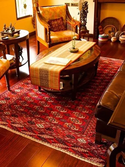 The living room at the Roopa Hariprasad Residence is furnished with classic wooden pieces and a rich, patterned rug over hardwood flooring. The entire composition evokes a sense of heritage and timeless comfort.