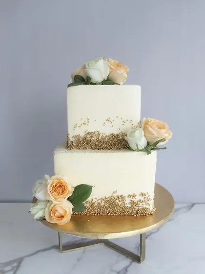 A modern and elegant two-tiered square wedding cake. The clean lines are softened by clusters of fresh peach and white roses and a delicate scattering of golden sugar pearls.