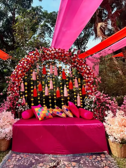 A lush, outdoor Mehendi setup with a floral arch in shades of pink and red, complemented by overhead drapes that add a canopy of color.