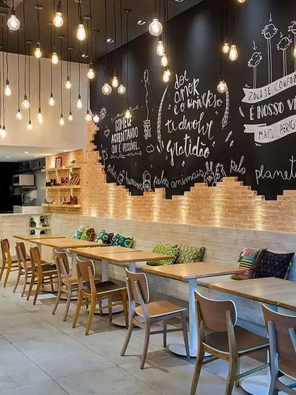 For this cafe, we created a warm, industrial-chic vibe using an exposed brick feature wall with custom chalkboard art. The space is lit with hanging Edison bulbs, and the seating arrangement with simple wooden tables and chairs provides a cozy, rustic setting for patrons.