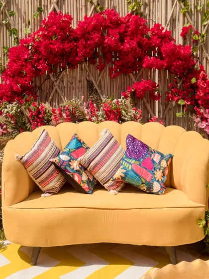 A close-up of the sunshine-yellow sofa, adorned with colorful and patterned cushions, set against the bougainvillea arch.