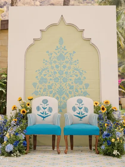 The main seating for the Haldi ceremony, featuring custom-printed chairs set against a backdrop with traditional Indian floral motifs. The blue and yellow color scheme is a modern twist on Haldi decor.