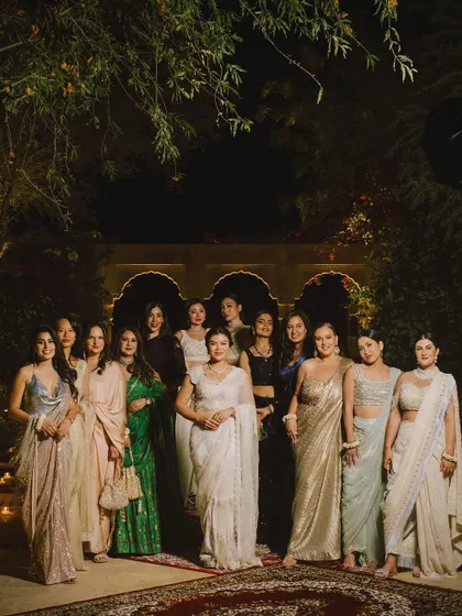 The bride and her squad, all looking regal in beautiful sarees and lehengas, pose for a group photo in a candlelit courtyard.