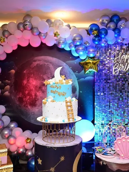 A spectacular moon-themed birthday party. The decor features a large moon backdrop, a silver sequin wall, and a balloon arch in shades of blue and silver, creating a celestial vibe.