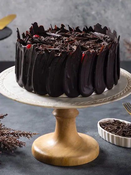 A black forest cake with intricate chocolate shavings. The lighting is designed to catch the glossy finish and sharp edges of the chocolate.