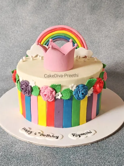 A vibrant rainbow-themed cake perfect for a young child's birthday. It features colorful fondant stripes, piped buttercream flowers, and a cute rainbow and crown topper.