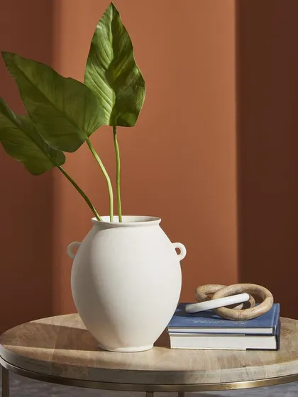 A large white vase styled with green leaves on a wooden side table. This in-context shot shows the product in a realistic home setting, helping customers visualize its scale and use.