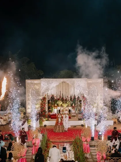 A wide shot of the Varmala ceremony on a grand stage, complete with dramatic lighting and pyrotechnics. I capture the full scale of your celebration.
