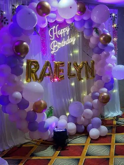 A stylish birthday decoration with a purple, white, and gold balloon ring. The custom name in gold balloons and the neon sign make it very personalized.