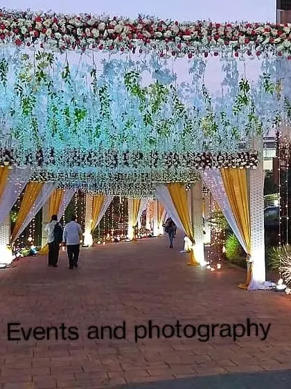 A bright and welcoming entrance for a daytime or evening event. The walkway is lined with yellow and white drapes, fairy lights, and a ceiling of hanging white flowers.