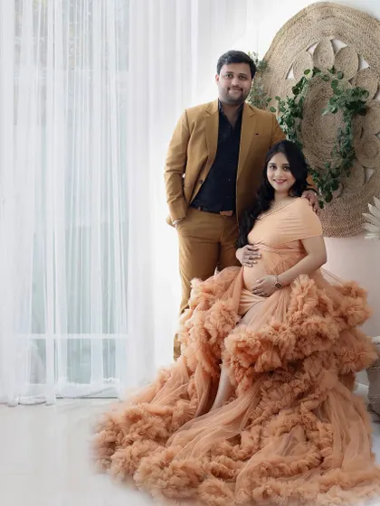 This image showcases a beautiful ruffled gown in a soft peach tone. The couple stands together in a light-filled studio space, creating a warm and elegant maternity portrait.