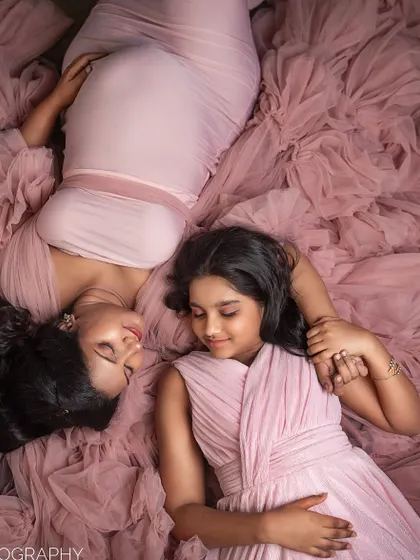 In a beautifully styled studio setting, a mother-to-be and her daughter lie together on a bed of soft pink tulle, sharing a quiet, loving moment. This image highlights the tender bond between a mother and her child as they await a new arrival.