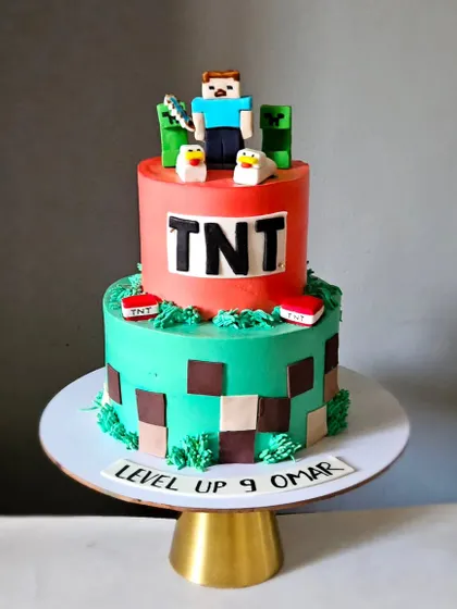 A two-tiered Minecraft cake, featuring a TNT block top tier and a pixelated bottom tier. The cake is topped with a Steve figurine and Creepers.