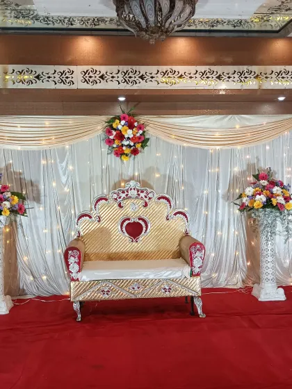 Here is a classic and elegant stage decoration for a formal function. We used cream-colored drapes with fairy lights, a red carpet, and a traditional golden throne sofa, accented with four colorful floral bouquets on pillars.