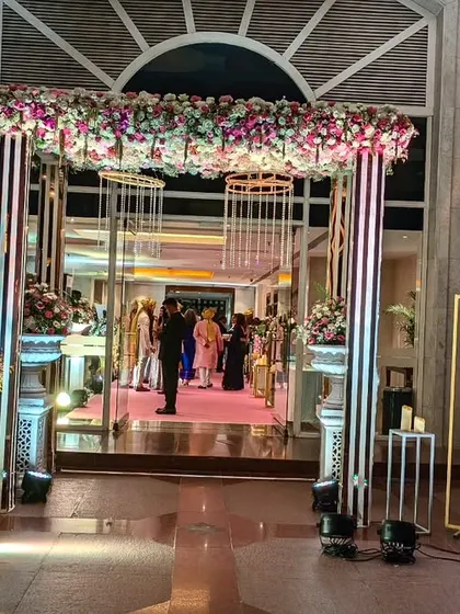 A grand entrance at The Lalit, featuring a modern structure with hanging crystals and lush floral arrangements. The couple's photos on easels add a personal touch to this glamorous welcome.