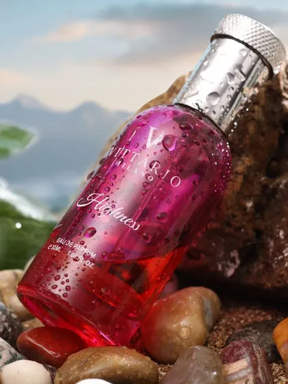 A pink perfume bottle rests on rocks with water droplets, suggesting a fresh, aquatic, and vibrant fragrance.