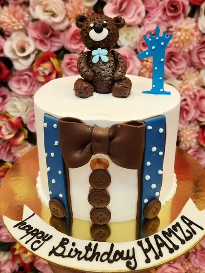 A charming teddy bear cake for a first birthday, designed to look like the bear is wearing suspenders and a bow tie.