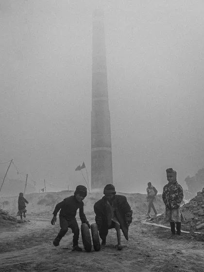 Children play with a tire in the foggy, industrial landscape of a brick kiln. This poignant black and white photo captures a moment of childhood play against a bleak backdrop.