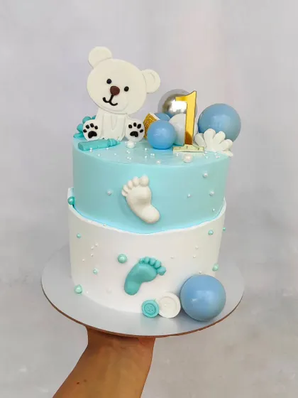 A sweet two-tier fault line cake for a first birthday. The design includes a cute teddy bear, baby footprints, and shades of blue.