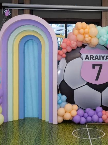 A full view of the sports-themed decor, with a faux grass floor and a large, personalized soccer ball backdrop surrounded by colorful pastel balloons.