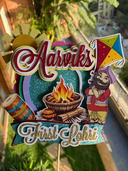 Celebrate a baby's first Lohri with this vibrant and festive handmade topper. It features traditional elements like a bonfire, dhol, and kite, all crafted from colorful and glittery cardstock.
