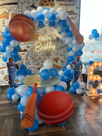 A cricket-themed 6th birthday party for a young sports fan. The setup includes giant cricket bat and ball props, player cutouts, and a blue and white balloon arch.