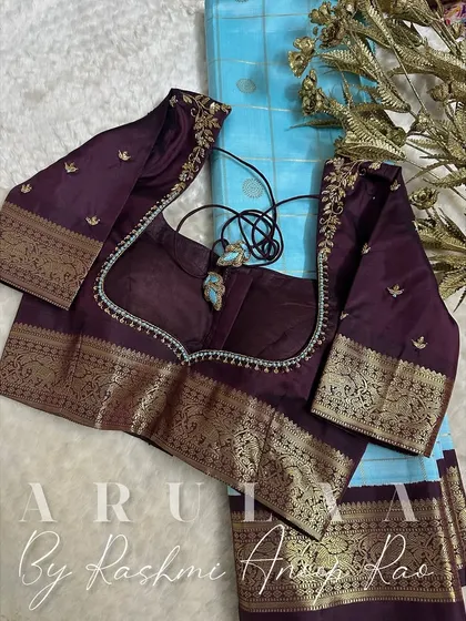 A deep wine-colored blouse paired with a light blue saree. The blouse features delicate floral vine embroidery on the shoulders and a simple beaded border.