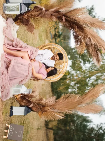 A rotated image of a couple's outdoor maternity shoot, featuring a wicker chair and pampas grass setup. The angle gives it a creative, dynamic feel.