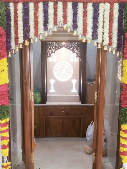 A pooja room entrance decorated with colorful vertical flower garlands and small bells. This frames the sacred space beautifully.