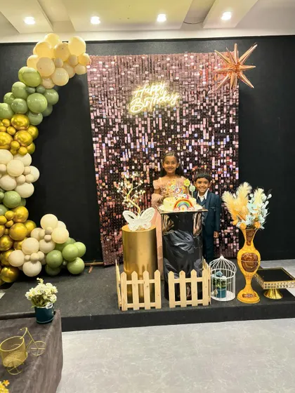 A stylish party setup with an olive green and gold balloon arch against a rose gold sequin wall. The children standing by the cake table show how these decorations create the perfect backdrop for birthday photos.